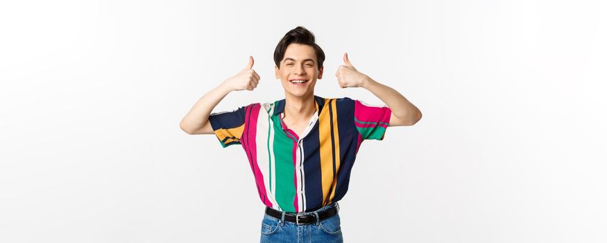 Young cheerful man smiling, showing thumbs up in approval, praise something good, standing over white background