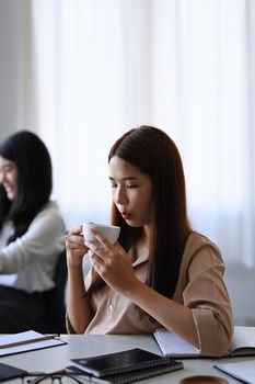 Peaceful young businesswoman sitting in office and drinking to coffee.