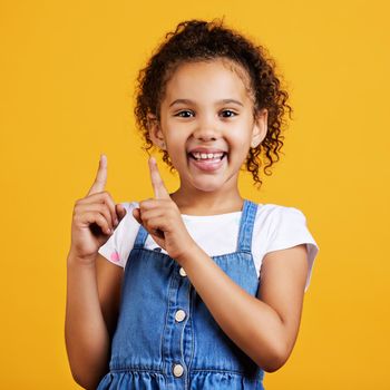 Studio portrait mixed race girl pointing upwards towards copyspace isolated against a yellow background. Cute hispanic child posing inside. Happy and cute kid showing or endorsing a company or product