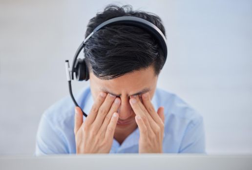 Stressed young asian male call centre agent getting a headache while working on computer in office. Consultant making mistakes and struggling with difficult customers when operating a support helpdesk
