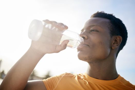 A jogger drinking a bottle of water in the sunlight. An active, healthy African American man quenching his thirst after exercising