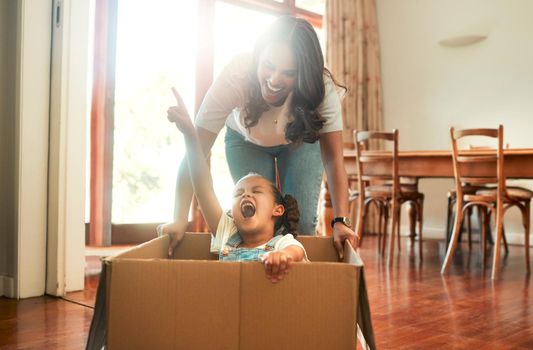 Mother and daughter playing with a cardboard box. Excited little girl sitting in a box. Parent pushing her daughter in a box. Parent having fun with her child at home. Cheerful mother and daughter