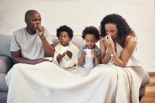 African american family of four feeling sick and unwell with flu and cold while covered in a blanket on a couch. Parents and two kids coughing and blowing nose from seasonal allergy symptoms and covid