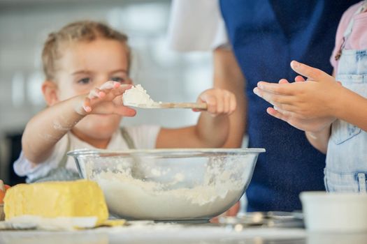 Little girl holding a spoon of flour. Family baking together at home. Two sisters baking with a parent. Caucasian girl putting flour into a bowl. Mother baking with her daughters. Siblings baking .