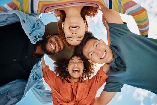 Group of diverse friends standing in a huddle against a blue sky. Multi-racial friends standing together arms around each other and smiling while looking down at the camera