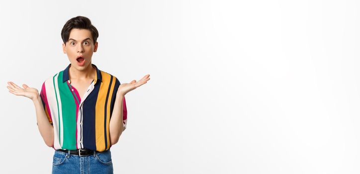 Image of surprised young male model staring at camera, spread hands sideways in complete disbelief, standing over white background