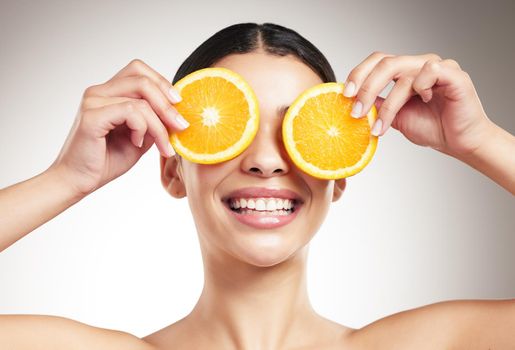 Young happy beautiful woman covering her eyes while holding an orange and posing against a grey studio background alone. One hispanic female smiling showing a fruit while standing against a background