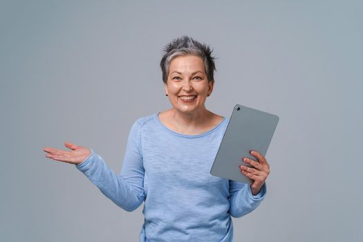 Happy middle age woman with grey hair and tablet pc in hands browsing, working, shopping online. Pretty woman in 50s wearing blue blouse isolated on grey. Mature people and technologies