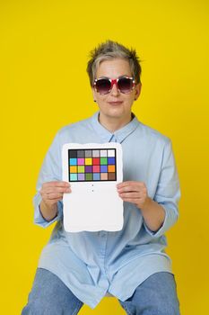 Sitting on a chair mature grey hair woman in UK flag sunglasses with color checker in hands looking at camera wearing blue shirt isolated on yellow background. Healthcare, aged beauty concept