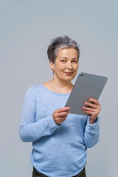 Grey haired woman with tablet pc in hands working online. Pretty woman in 50s wearing blue blouse isolated on grey. Mature people and technologies
