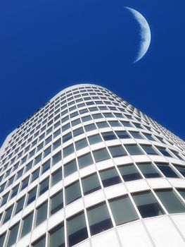 Half a moon over a building skyscraper, shining in bright blue sky. Below the crescent of quarter moon over a tall modern structural architecture, foreboding doomed capitalism or an economical crisis