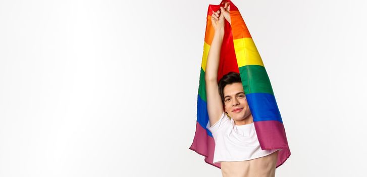 Vertical view of beautiful androgynous gay man raising rainbow flag and smiling happy, standing in crop top and jeans against white background