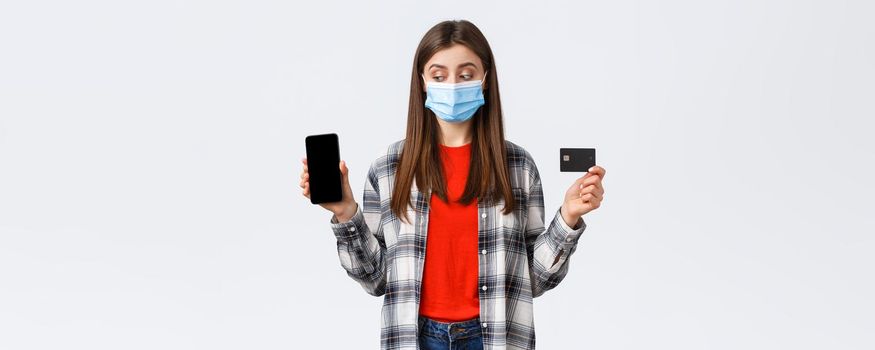 Coronavirus outbreak, working from home, online shopping and contactless payment concept. Girl in medical mask showing mobile phone and credit card, peek at screen making order