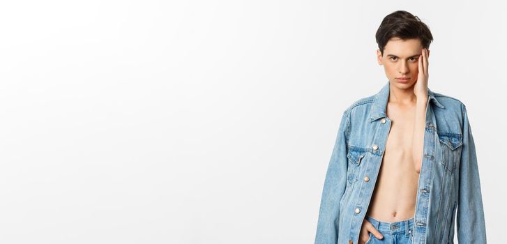Handsome and sassy gay man wearing denim jacket on bare torso, touching his face and looking confident at camera, standing over white background
