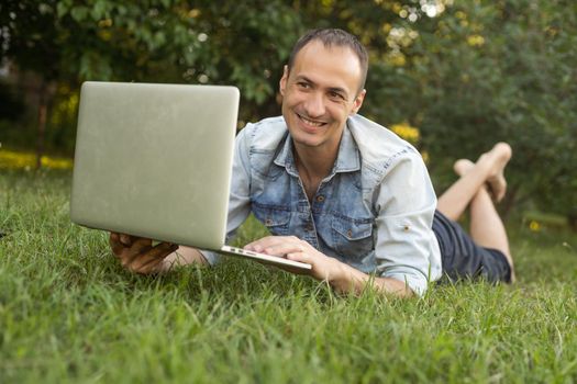 caucasian hispanic man using headphones and sitting on grass working with laptop. Casual man working outdoors