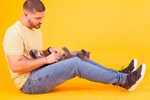 Young man rubbing his cat belly in studio over yellow background
