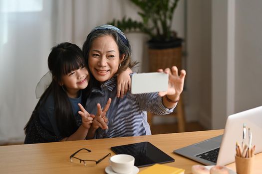 Happy asian grandma making selfie with cute little granddaughter, enjoying carefree leisure weekend activity together in living room