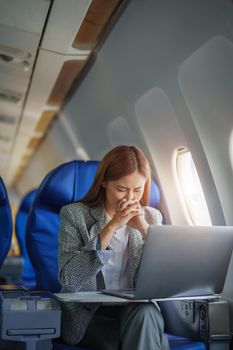 portrait of a successful Asian businesswoman or entrepreneur in a formal suit on an airplane seated in Business Class shows a thoughtful and stressed face with using laptop during the flight