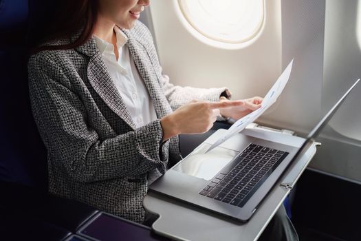 A successful asian businesswoman or female entrepreneur in formal suit in a plane sits in a business class seat and uses a documents with computer laptop during flight