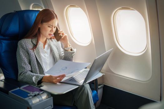 Portrait of a successful Asian businesswoman or entrepreneur in a formal suit on an airplane sitting in business class using a phone, computer laptop with a serious expression during the flight