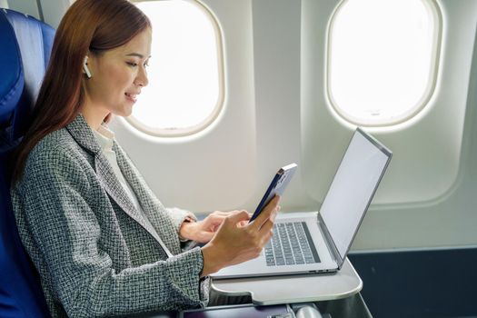 A successful asian businesswoman or female entrepreneur in formal suit in a plane sits in a business class's seat and uses a smartphone during flight