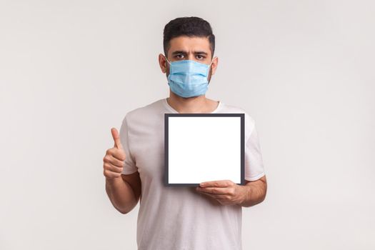 Man in hygienic mask showing thumb up and holding white poster, mockup template