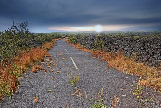 Empty road through a field with burnt grass and cloudy sky with copy space. A curved countryside road or open asphalt roadway between dry land near Mauna Kea volcanic mountainside, Hawaii, Big Island