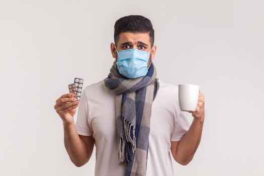 Portrait of scared man in protective hygienic mask holding pills and tea cup, looking desperate