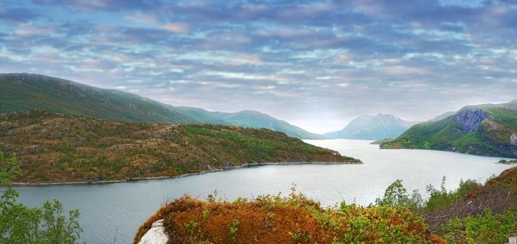 Landscape of a river between hills and mountains. Green foliage by the riverbank with a blue sky in Norway. Calm water near a vibrant wilderness against a bright cloudy horizon. Peaceful nature scene