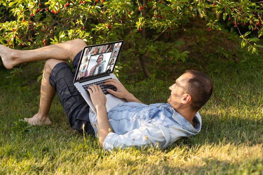 Freelancer man with laptop working in garden sit in chair on grass outdoors. Young blogger male work on computer in public park processes video for social media content. Place of work, distance job