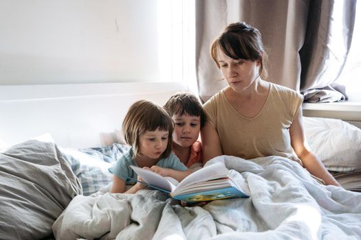 Woman reading a book to siblings while they are all sitting in the bed