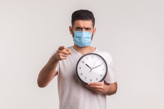 Time to be cautious. Man in protective hygienic mask holding clock, warning of novel virus epidemic