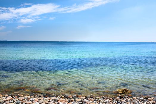 Calm ocean waves washing onto stones at an empty beach shore with sailboats cruising in the horizon. Scenic landscape for a relaxing summer vacation. Copy space at the sea with a blue sky background