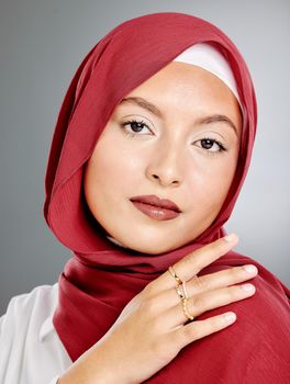 Portrait of stylish muslim woman with makeup and gold rings isolated against grey background. Edgy young hijab lady feeling confident in her red scarf and trendy costume jewelry. Modesty and beauty