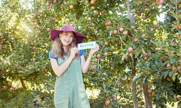 Female farmer recruiting workers to help with her startup. Happy young woman holding a hiring sign on a farm close to apple orchards. Farm labor shortage, agriculture job market and employment