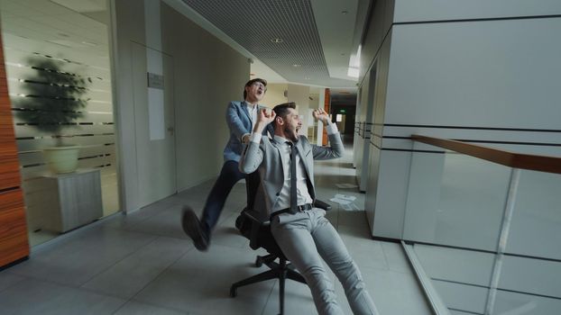 Two crazy businessmen riding office chair and throwing papers up while having fun in lobby of modern business center