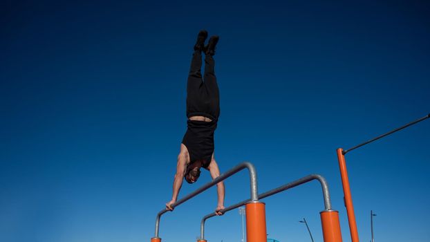 Shirtless man doing handstand on parallel bars at sports ground.
