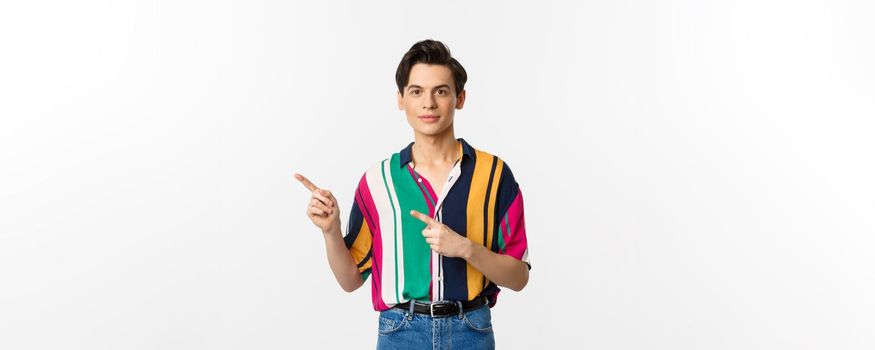 Image of stylish androgynous man smiling, pointing fingers left at logo, showing promo offer, standing over white background