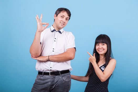 Smiling asian girlfriend pointing at his caucasian boyfriend while he shows ok hand gesture.