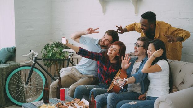Group of joyful friends taking selfie photos on smartphone camera while celebrating at party with beer and snacks at home indoors