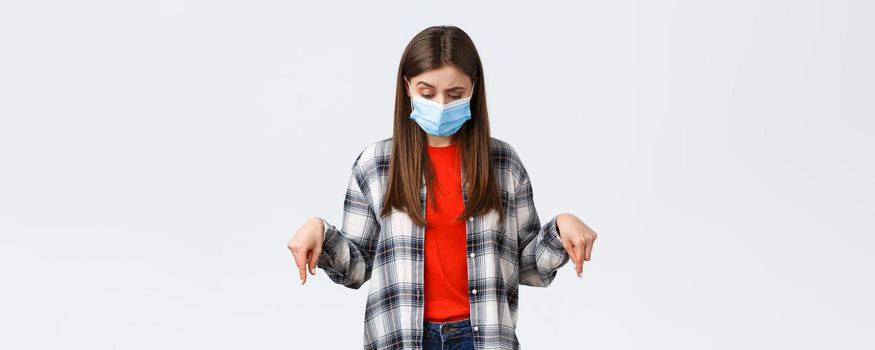 Coronavirus outbreak, leisure on quarantine, social distancing and emotions concept. Hesitant and unsure attractive woman in medical mask, looking down, pointing at strange thing