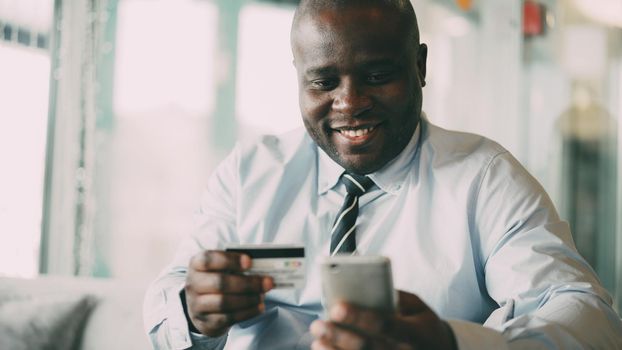 Cheery African American businessman in formal clothes paying online bill keeping credit card and smartphone in his hands in glassy cafe