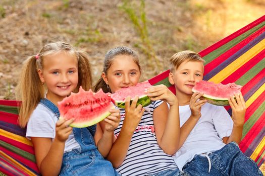 children eat watermelon and joke, outdoor, sitting on a colorful hammock.