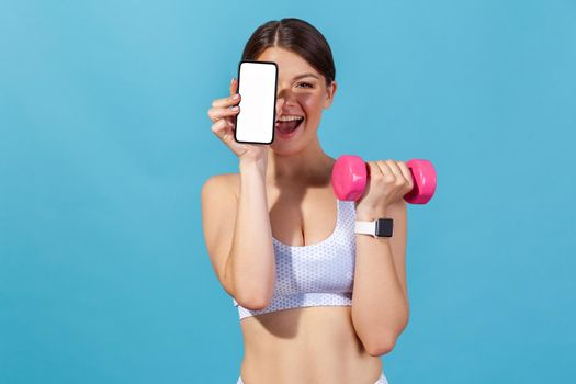 Happy athletic woman in white sports top holding empty screen smartphone and pink dumbbell, doing fitness pilates exercising watching tutorial on phone. Indoor studio shot isolated on blue background