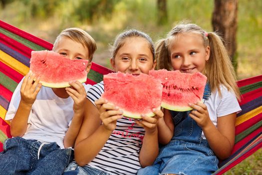 children eat watermelon and joke, outdoor, sitting on a colorful hammock.
