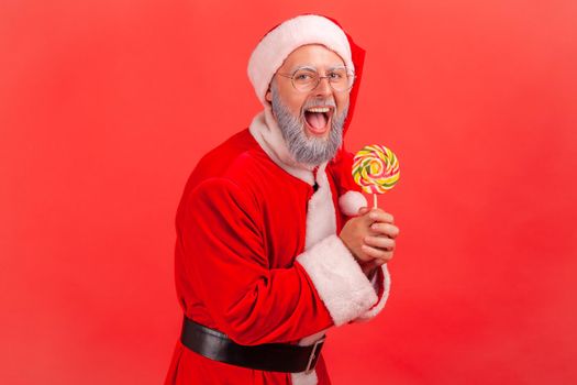 Portrait of happy amazed smiling elderly man with gray beard wearing santa claus costume standing with sugary lollypop in hands, looking at camera.