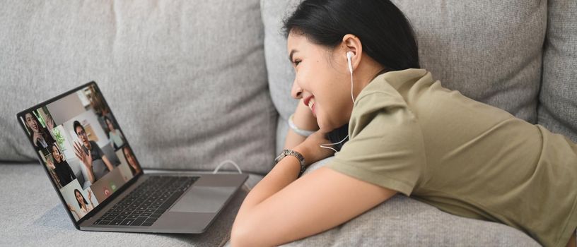 Cheerful young woman communicating by video conference via laptop while lying on couch at home