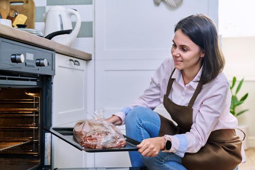 Young woman with meat on baking tray opening oven, kitchen at home