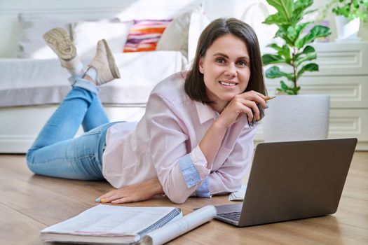 Attractive young woman looking at camera lying on floor at home with laptop