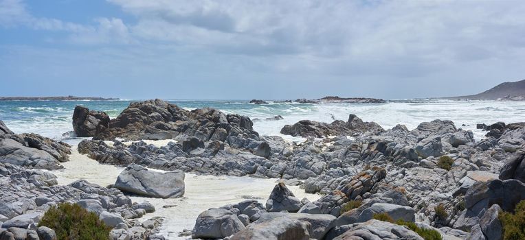 A rocky coastline at a beach in Cape Town with overcast clouds. A shore with rocks at the ocean with some waves. Rocky coast of mountain sea with on a cold day by a rock pool.